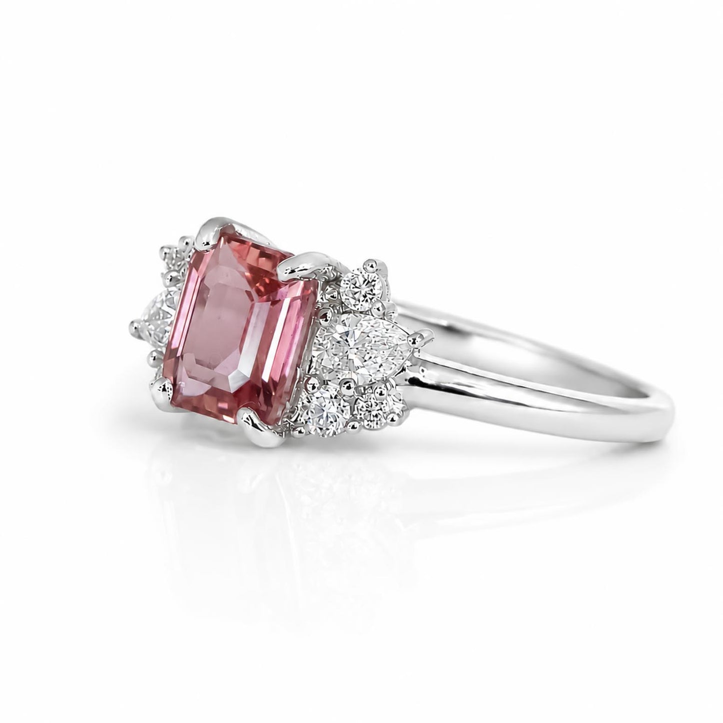 side view of a platinum ring showcasing an emerald cut padparadcsha sapphire with a cluster of diamonds on its side, photographed on a white background