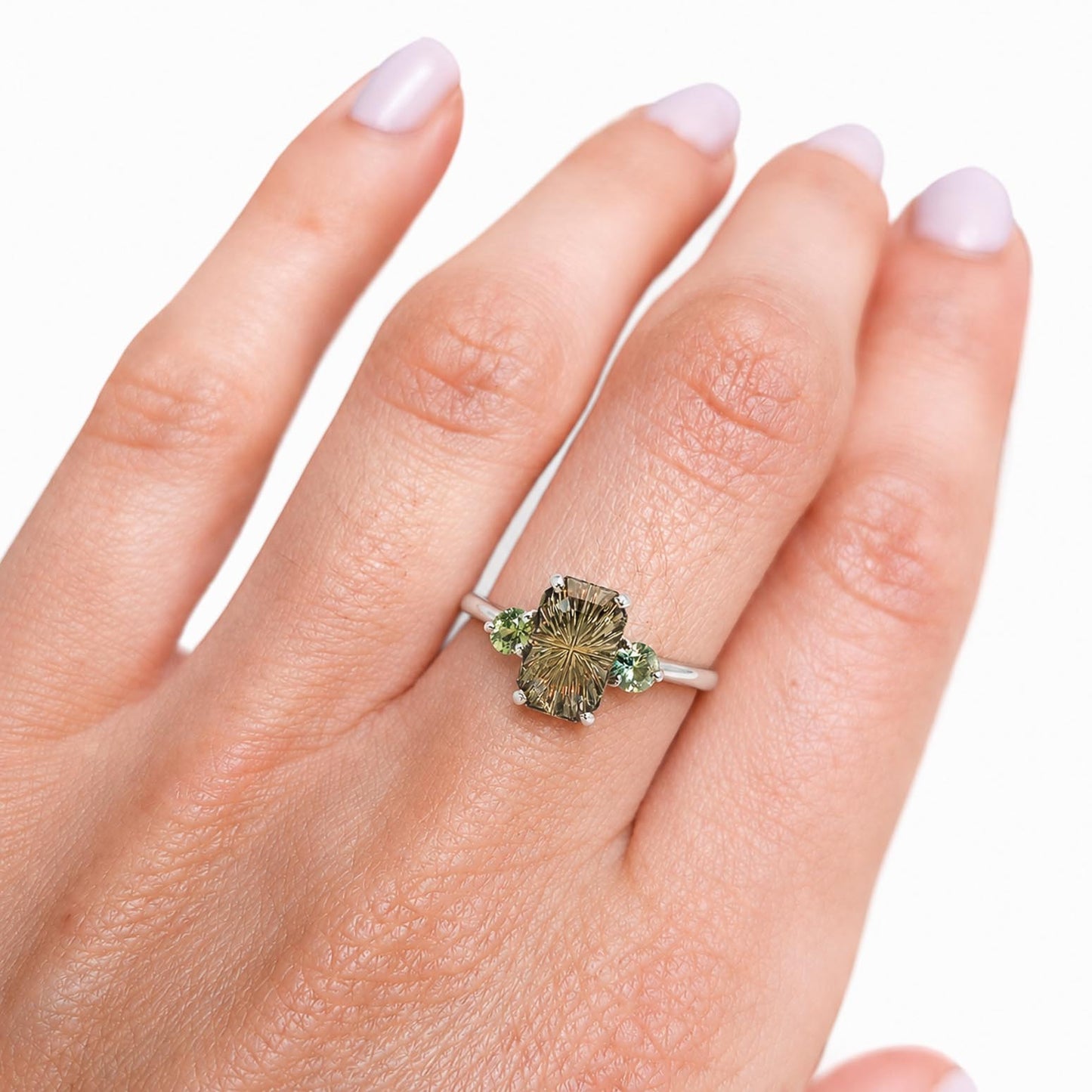 A close-up of a hand wearing a ring with a central elongated octagon-cut green sapphire and round teal green side Australian sapphires on a white gold band, set against a plain white background.