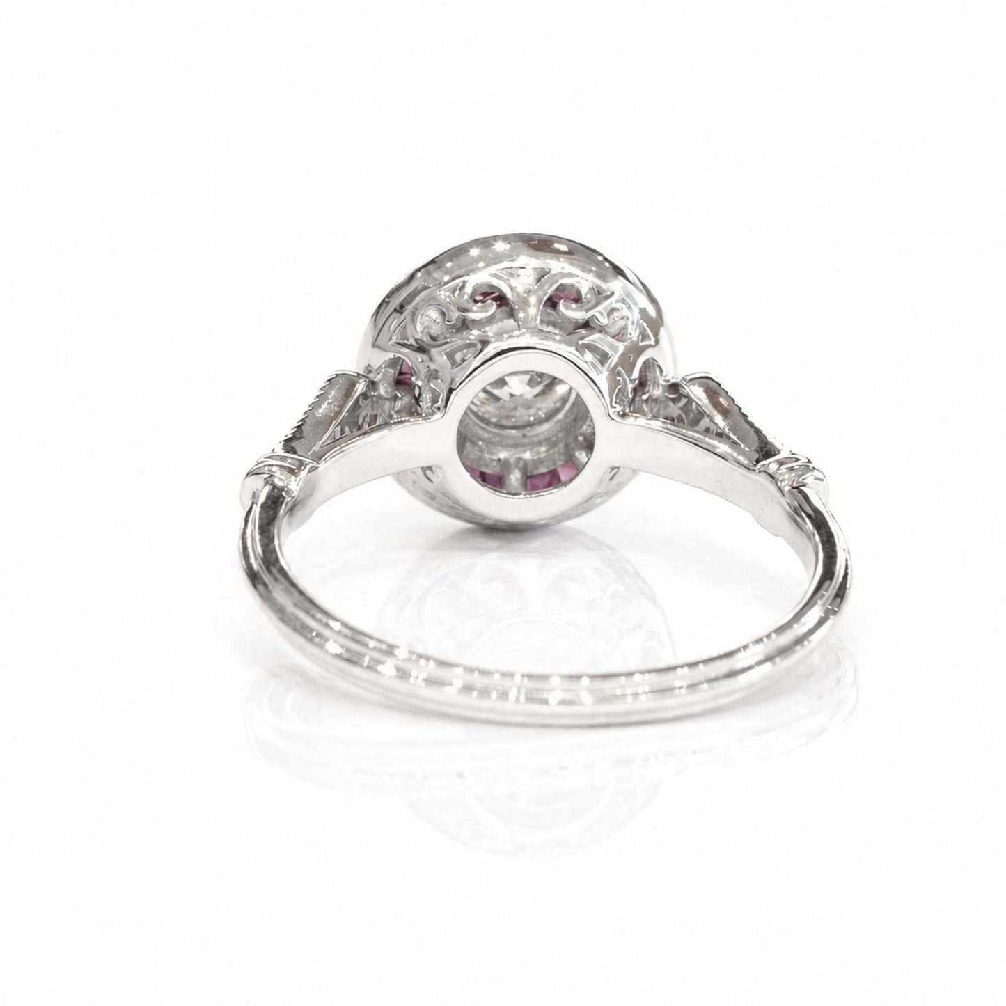 Back view of an antique-style white gold ring featuring a circular delailed basket with small rubies surrounding the centre diamond on a polished band, shown on a reflective white surface.