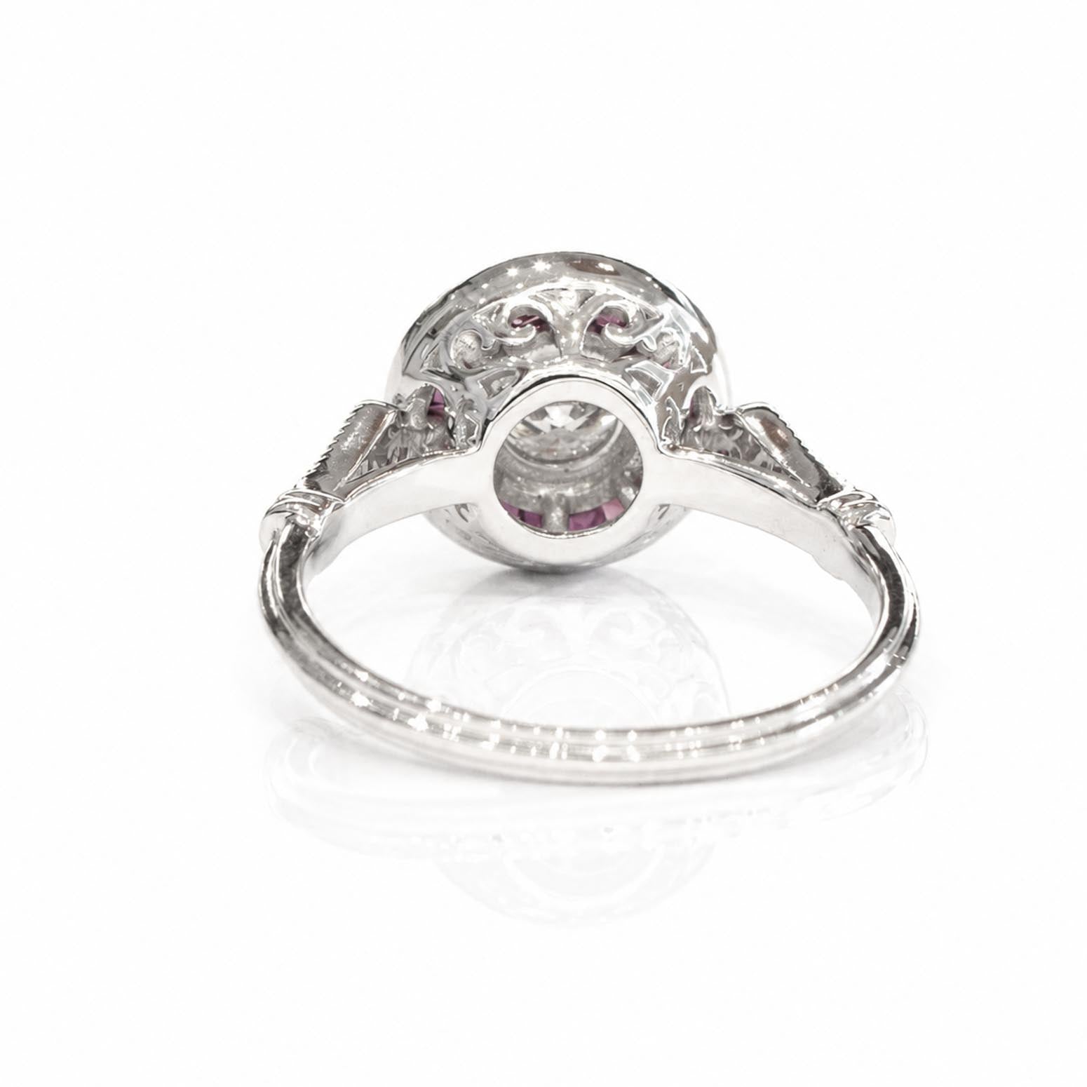Back view of an antique-style white gold ring featuring a circular delailed basket with small rubies surrounding the centre diamond on a polished band, shown on a reflective white surface.