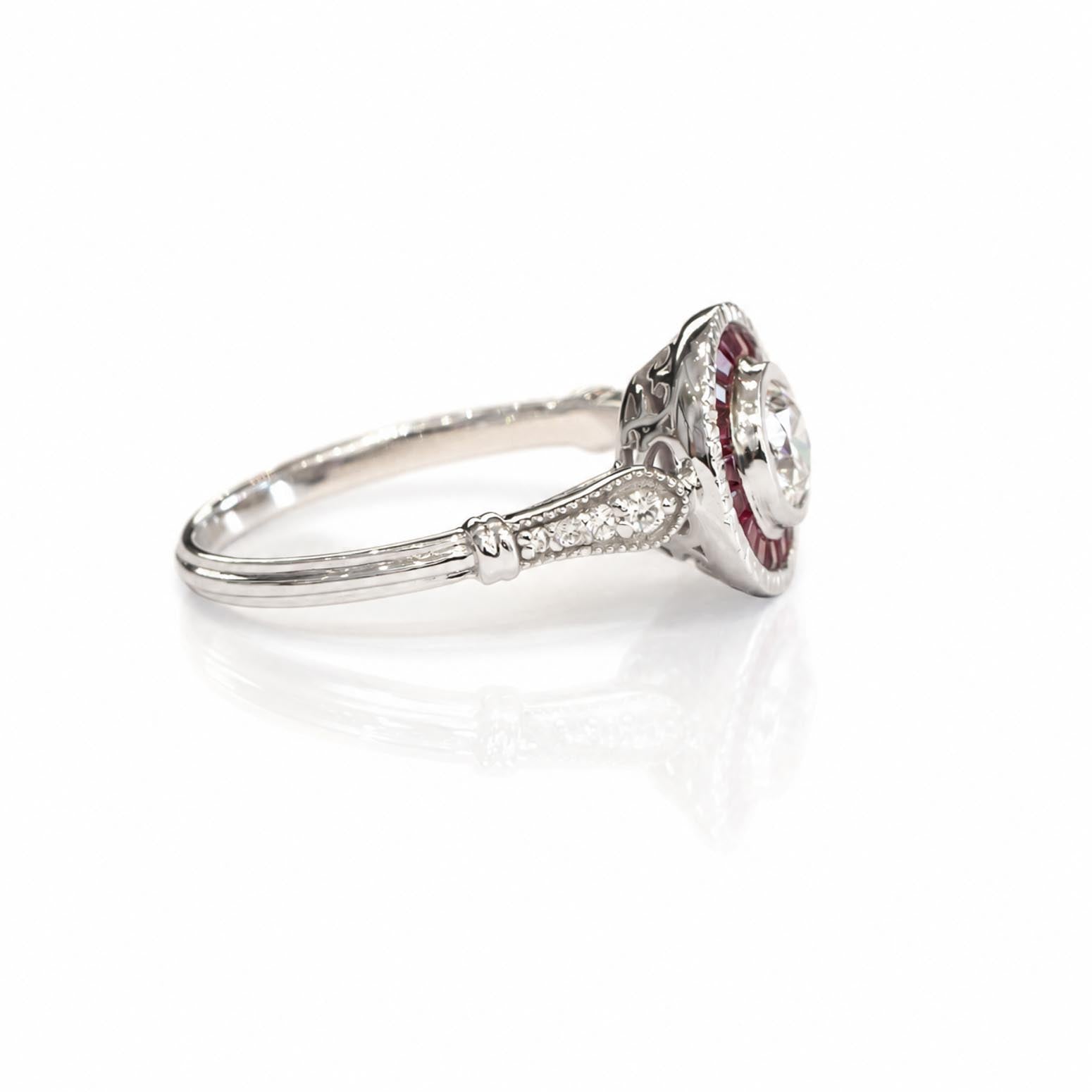 An antique-style white gold ring featuring a round diamond surrounded by a halo of custom cut baguette rubies and a row of accent diamonds on the band, shown on a reflective white surface.