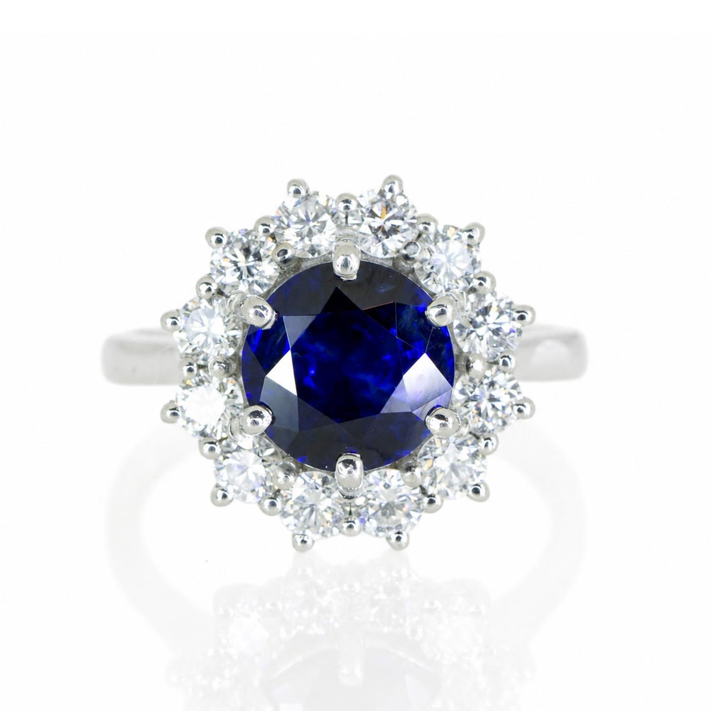 A platinum ring featuring a round deep blue sapphire center stone surrounded by a halo of round brilliant-cut diamonds, set against a clean white background with a soft reflection