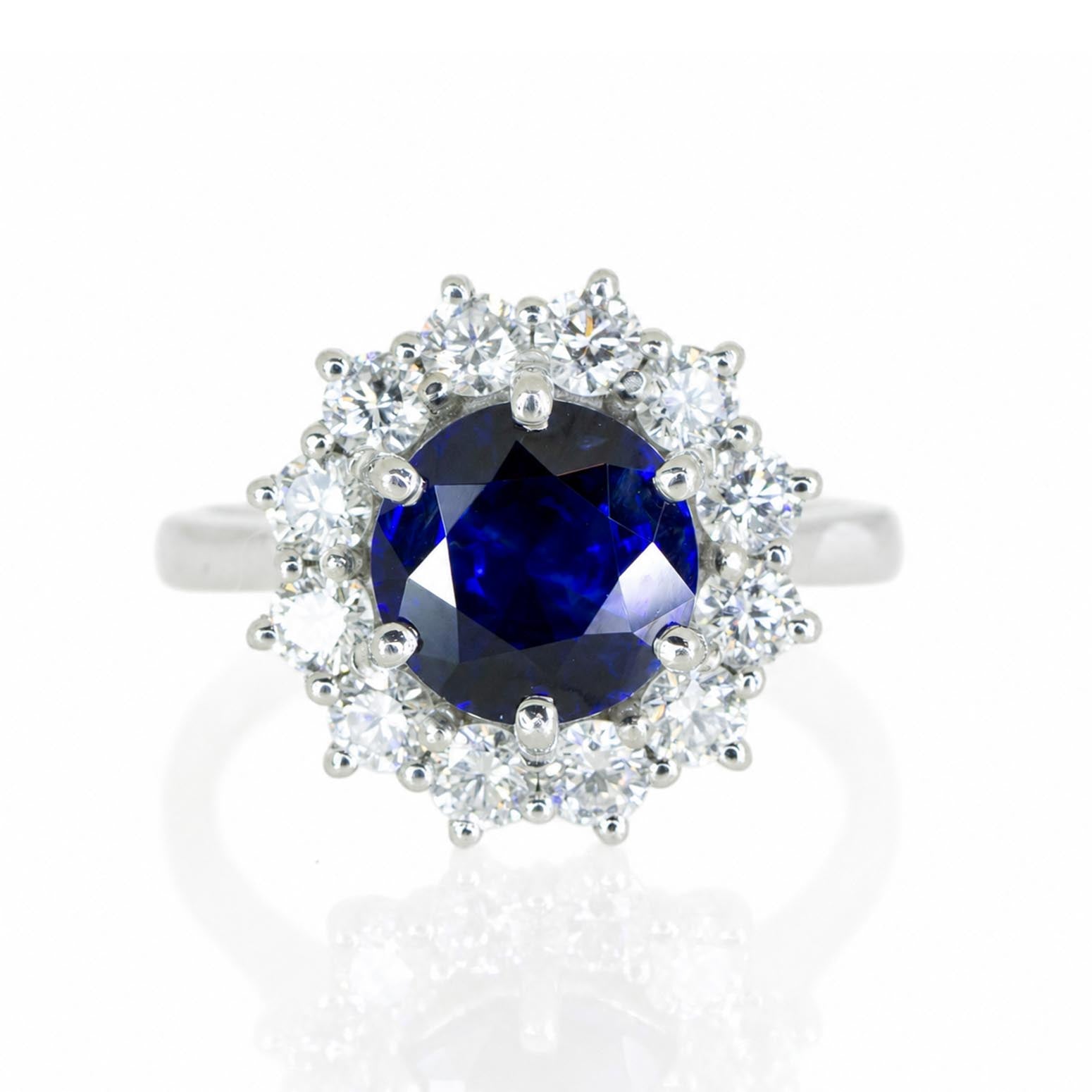 A platinum ring featuring a round deep blue sapphire center stone surrounded by a halo of round brilliant-cut diamonds, set against a clean white background with a soft reflection
