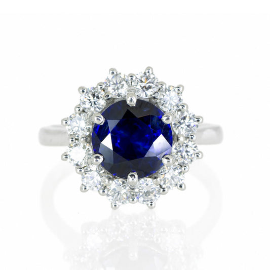 A platinum ring featuring a round deep blue sapphire center stone surrounded by a halo of round brilliant-cut diamonds, set against a clean white background with a soft reflection