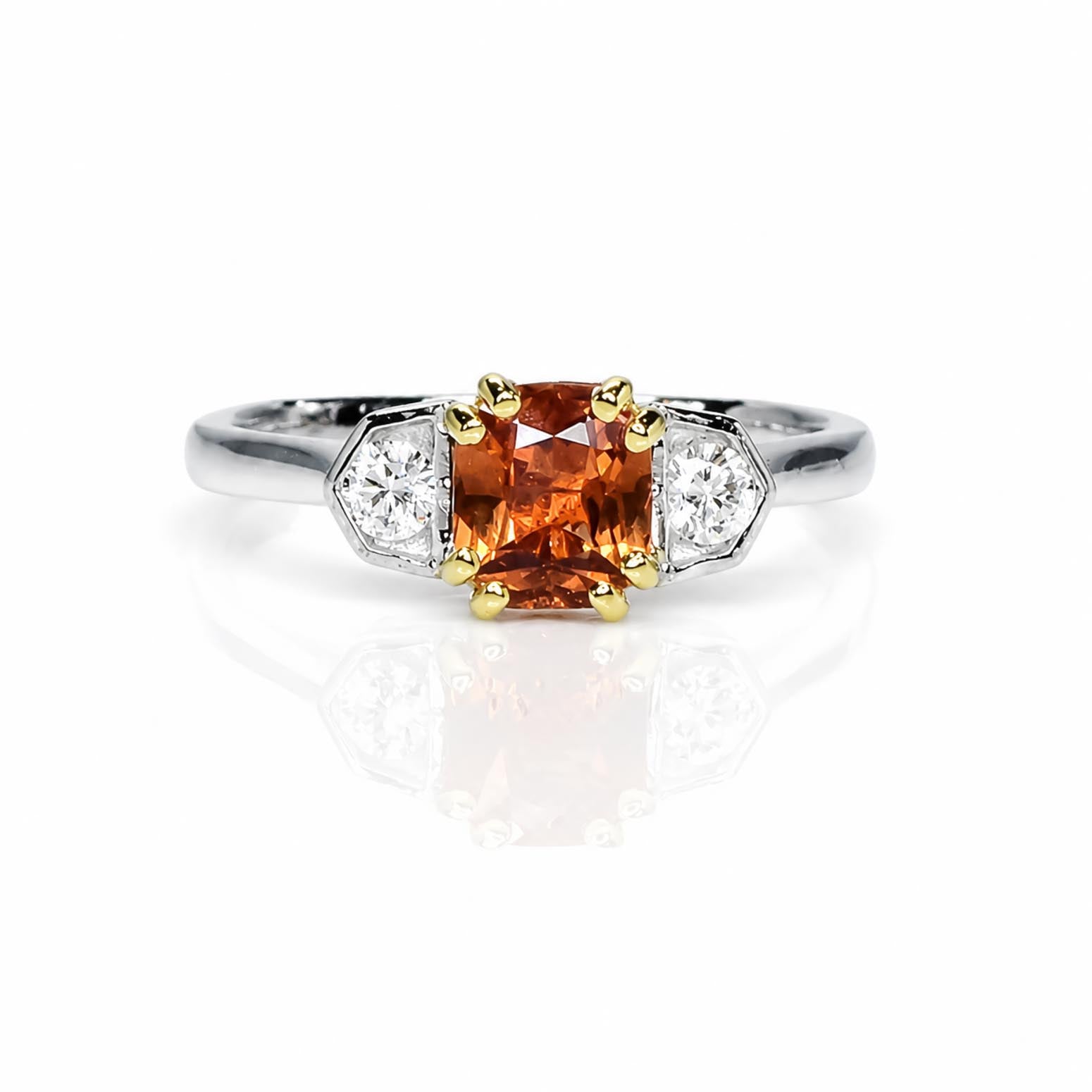 A three-stone diamond ring featuring a central padparadscha sapphire, flanked by two round-cut diamonds in platinum setting with yellow gold accents on the prongs, set against a white background with a soft reflection.