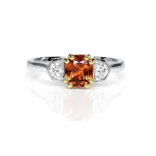 A three-stone diamond ring featuring a central padparadscha sapphire, flanked by two round-cut diamonds in platinum setting with yellow gold accents on the prongs, set against a white background with a soft reflection.