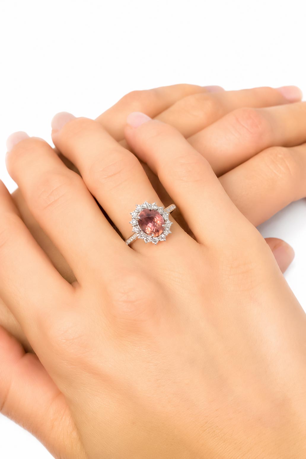 A close-up of a person's hands wearing a platinum ring with a natural padparadscha sapphire surrounded by diamonds, set against a plain white background.