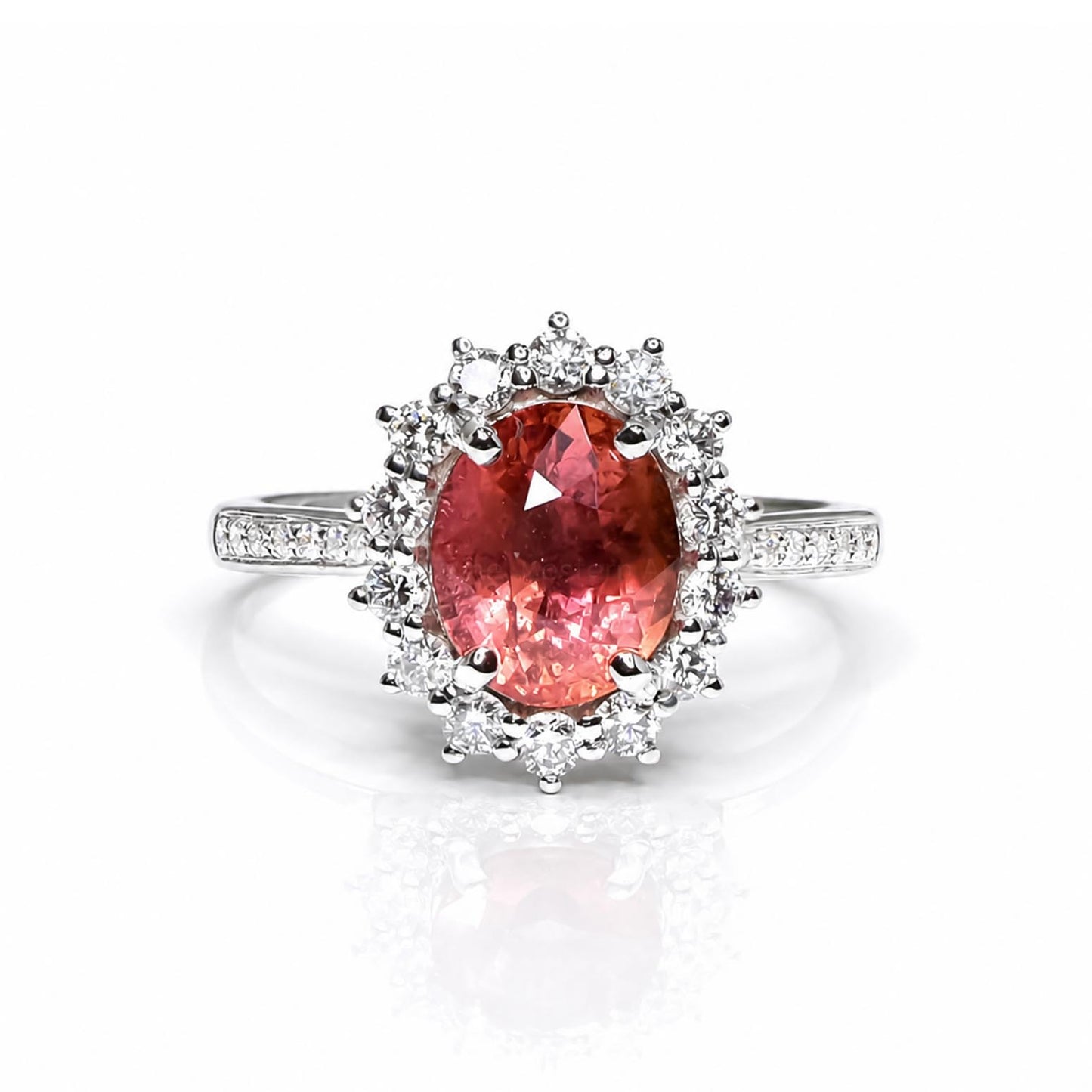 A platinum ring featuring a large oval pinkish-orange padparadcsha sapphire center stone surrounded by a halo of diamonds, with a pavé-set platinum band, shown on a white background with a soft reflection.