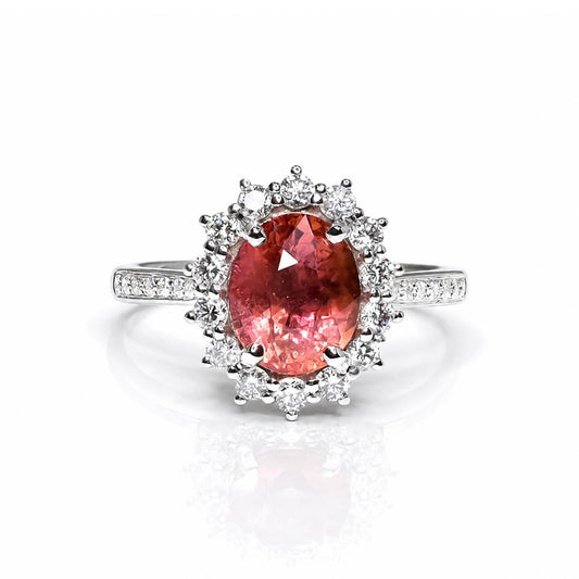 A platinum ring featuring a large oval pinkish-orange padparadcsha sapphire center stone surrounded by a halo of diamonds, with a pavé-set platinum band, shown on a white background with a soft reflection.