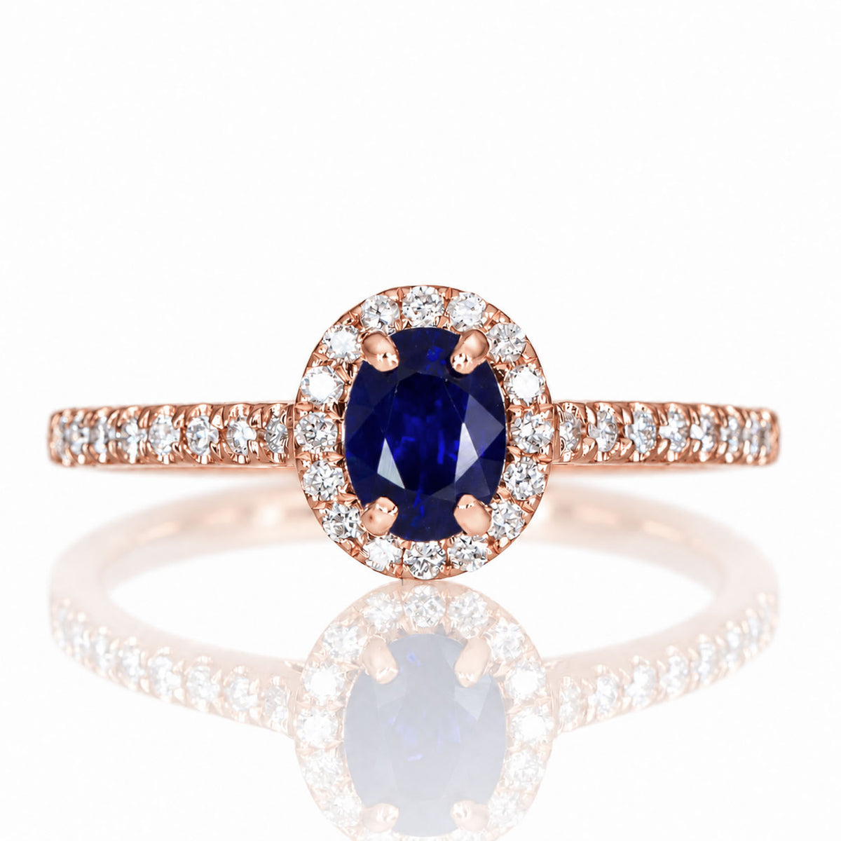 A rose gold ring with an oval cut, natural blue sapphire surrounded by a halo of diamonds, shown on a white background with a soft reflection