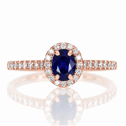 A rose gold ring with an oval cut, natural blue sapphire surrounded by a halo of diamonds, shown on a white background with a soft reflection