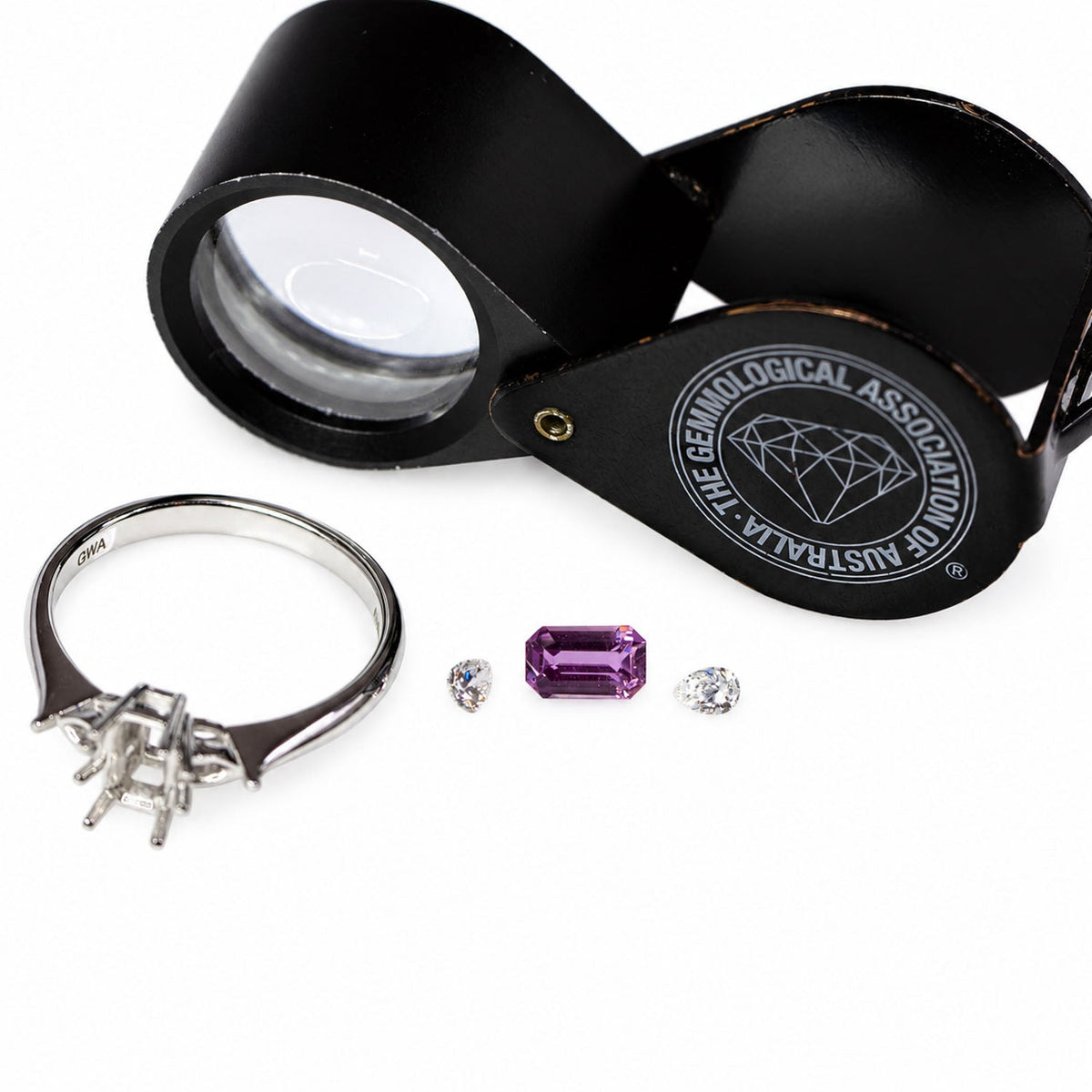 The pink sapphire, diamonds and white gold setting laid out beside a gemmologist's loupe