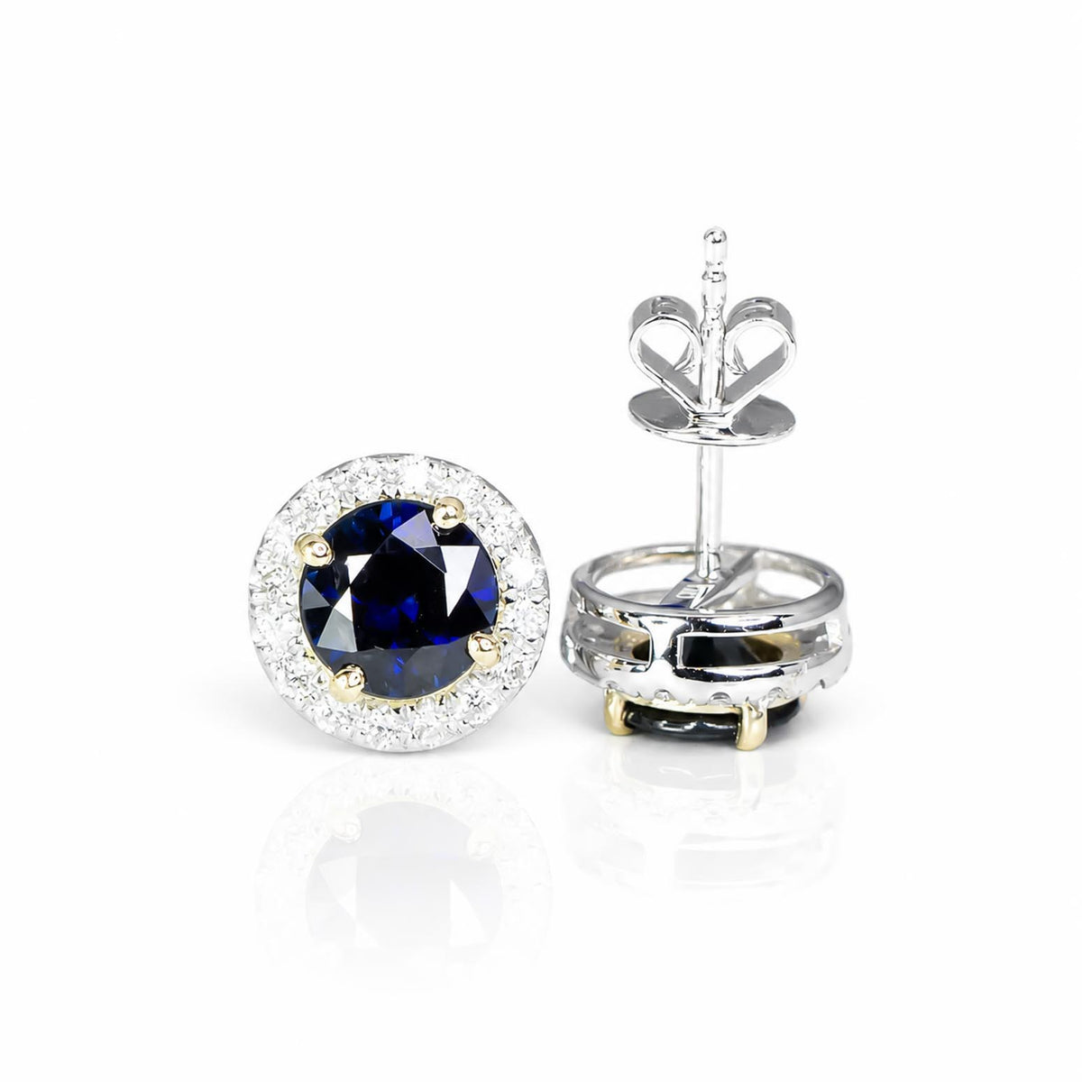 A pair of round stud earrings featuring a deep blue sapphire center stone surrounded by a halo of diamonds, with one earring shown from the front and the other from the side/back to display the post and butterfly back. The metal is white and yellow gold.