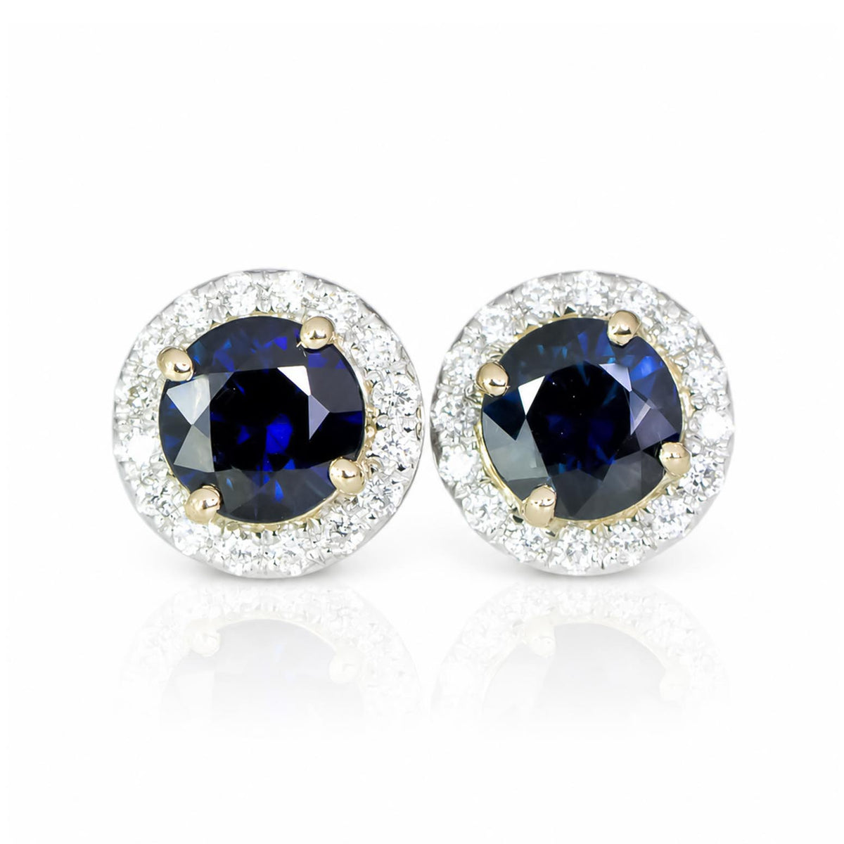 A pair of round stud earrings featuring deep blue sapphires surrounded by a halo of diamonds in a yellow gold and white gold setting, displayed on a clean white background with a soft reflection.