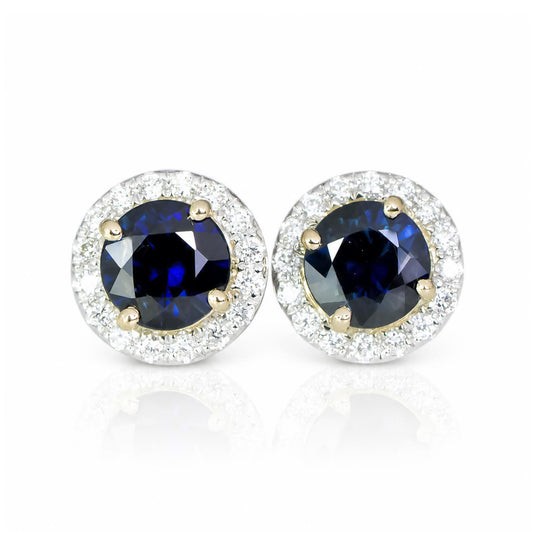 A pair of round stud earrings featuring deep blue sapphires surrounded by a halo of diamonds in a yellow gold and white gold setting, displayed on a clean white background with a soft reflection.