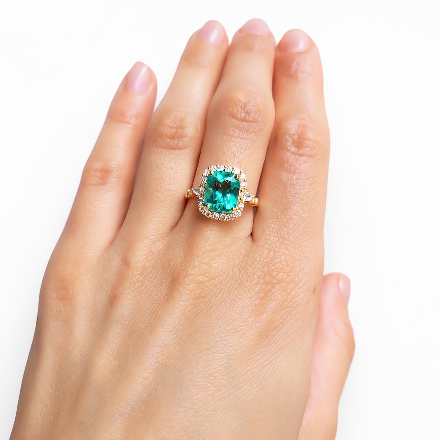 A gold ring featuring a cushion-cut Colombian emerald surrounded by a halo of small round brilliant-cut diamonds, shown worn on a hand against a plain white background.