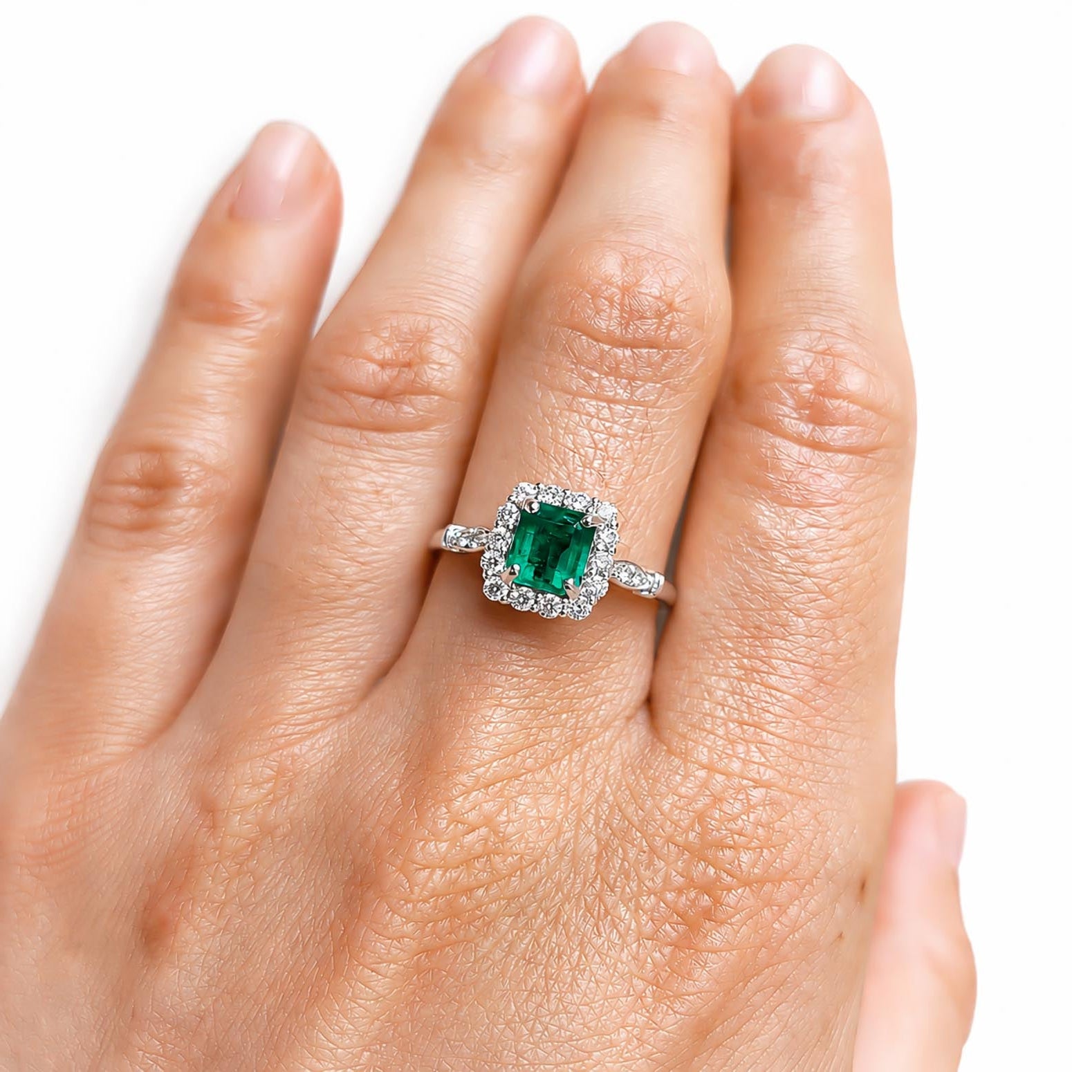A close-up of a hand wearing a platinum ring with an emerald-cut Colombian emerald centre stone and a halo of diamonds.