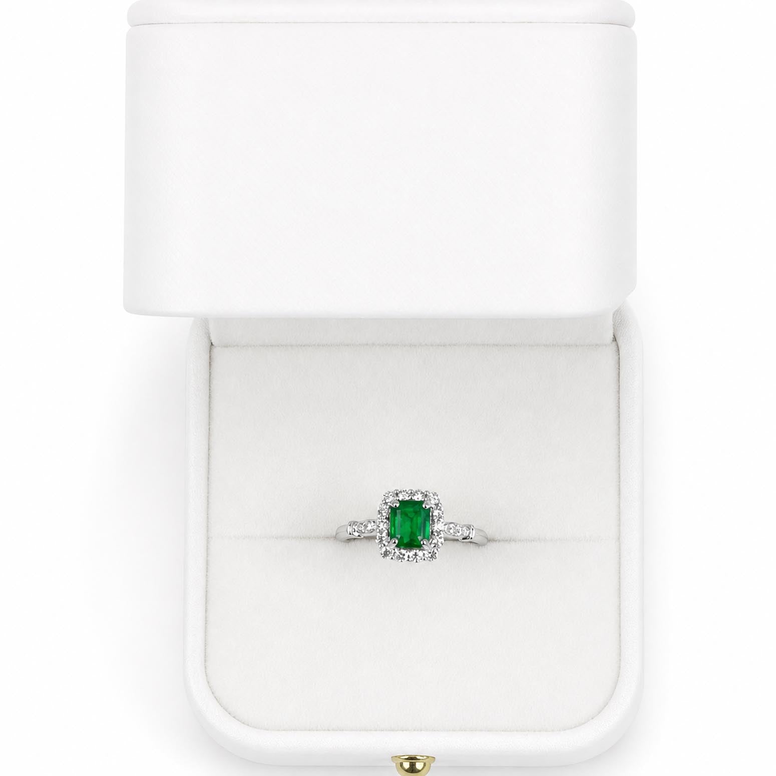 An emerald-cut Colombian emerald ring with a halo setting in platinum, displayed inside an open white jewellery box.