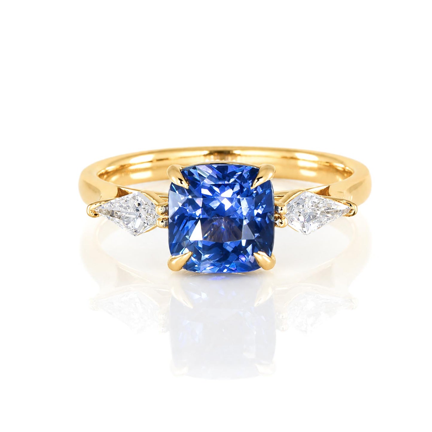 A yellow gold ring featuring a cushion-cut natural blue sapphire and natural kite-cut diamonds to its side on a polished band photographed on a white background 