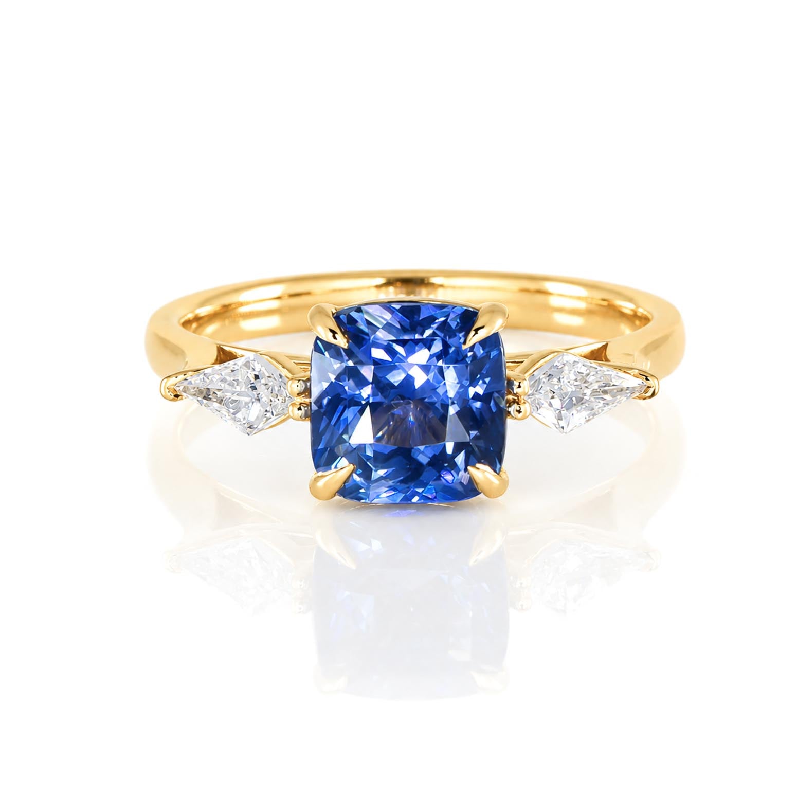 A yellow gold ring featuring a cushion-cut natural blue sapphire and natural kite-cut diamonds to its side on a polished band photographed on a white background 