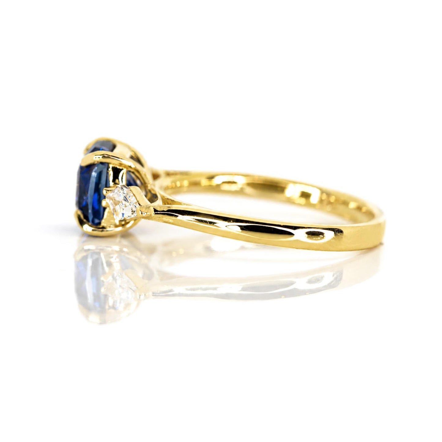 A yellow gold ring featuring a cushion-cut blue sapphire flanked by kite-cut diamonds, shown on a reflective white surface.