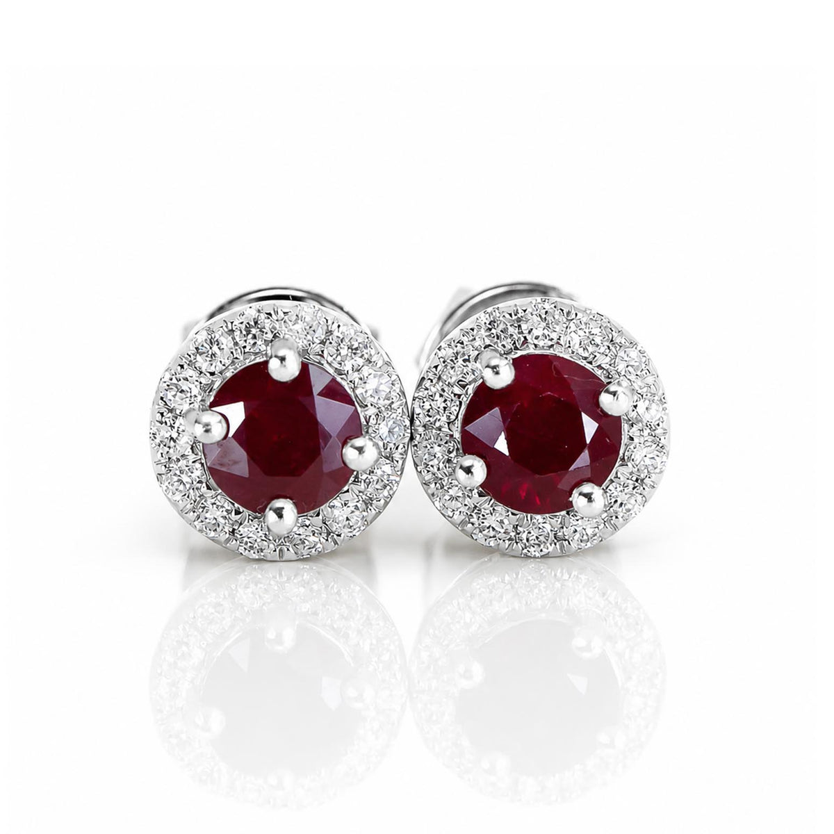 A pair of natural ruby earrings featuring round cut rubies surrounded by a halo of natural diamonds in 18K white gold shown on a white background 