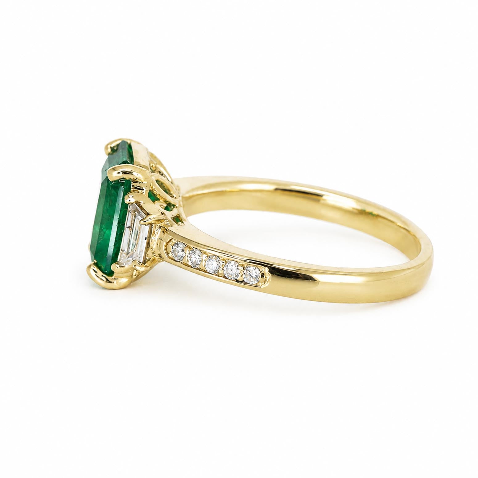 side view of a yellow gold ring featuring an emerald-cut Colombian emerald and baguette-cut and round diamonds on the shank, set against a plain white background.
