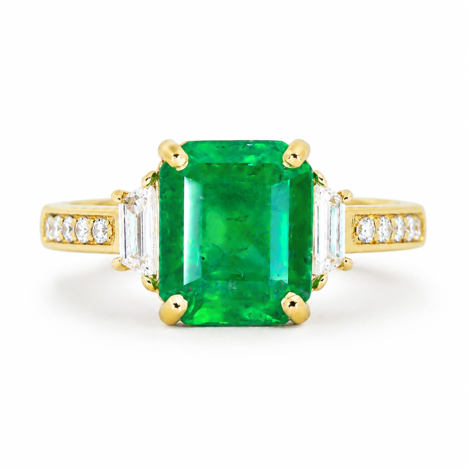 A yellow gold ring featuring a large emerald-cut Colombian emerald gemstone, flanked by baguette-cut side diamonds and a row of small round diamonds on the shoulders, set against a plain white background.