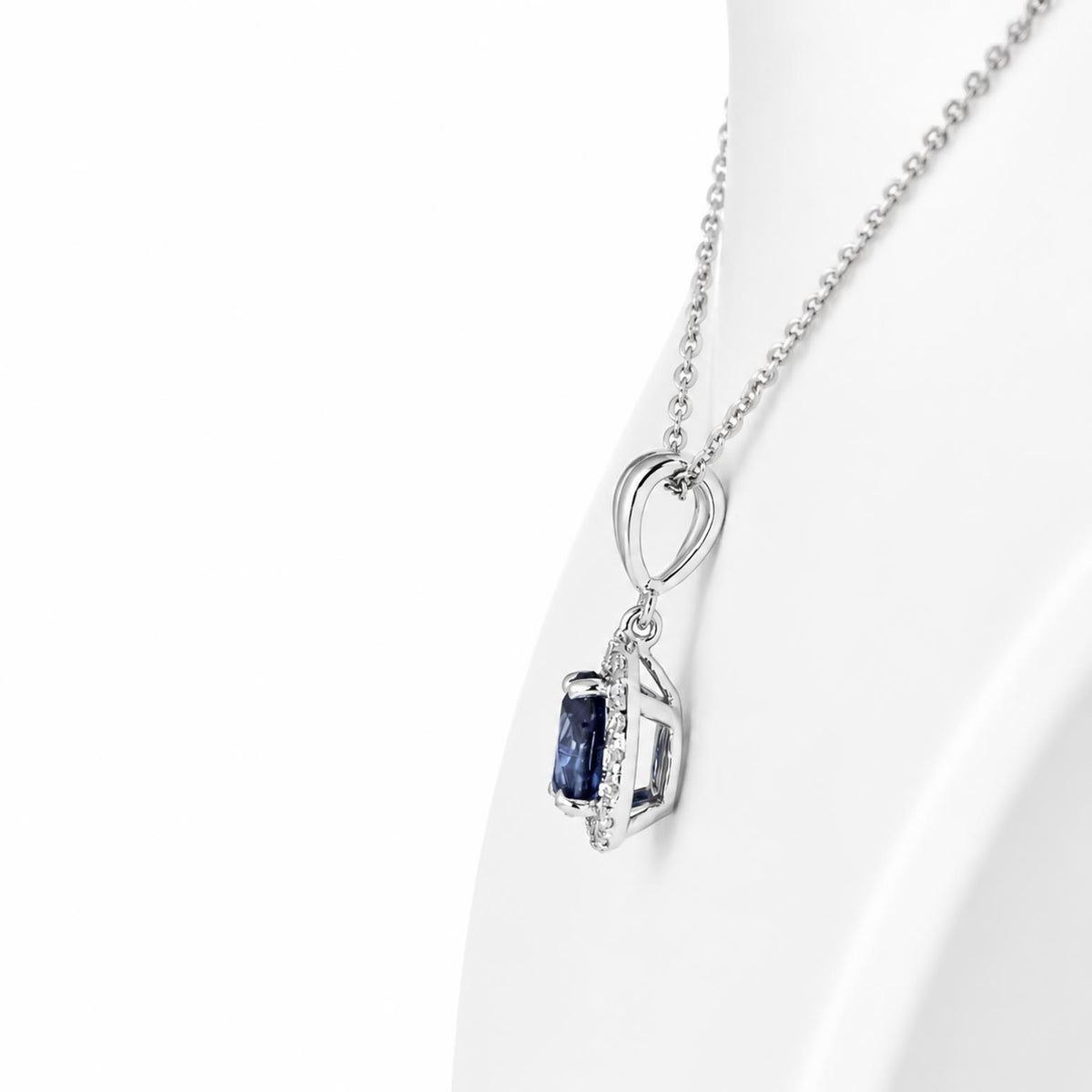 A white gold pendant necklace with a pear-cut blue sapphire and small accent diamonds, displayed on a white curved surface against a white background.