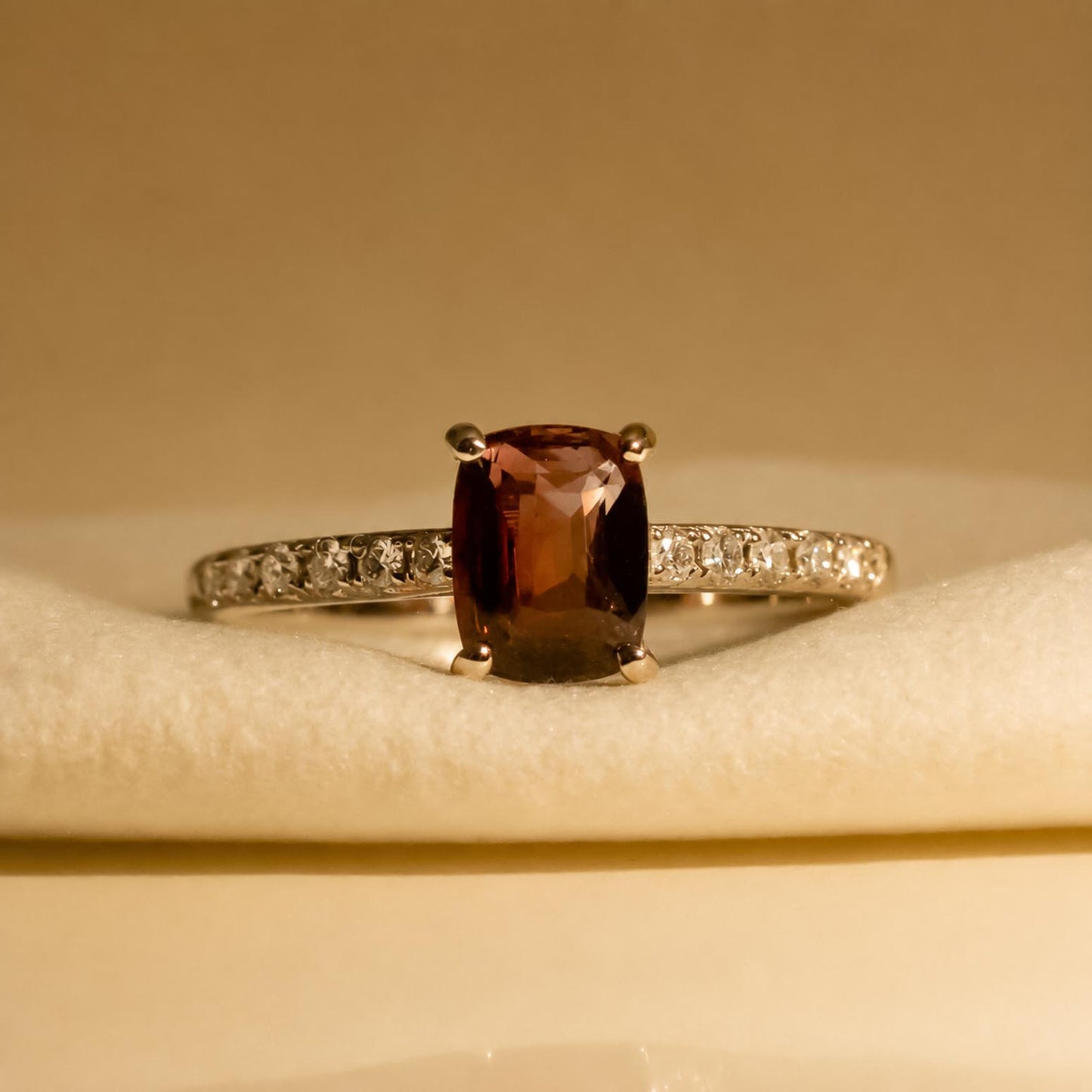 A white gold ring featuring a cushion-cut alexandrite center stone and a pavé diamond band, displayed on a soft cream-colored velvet surface and shown under incandescent light 
