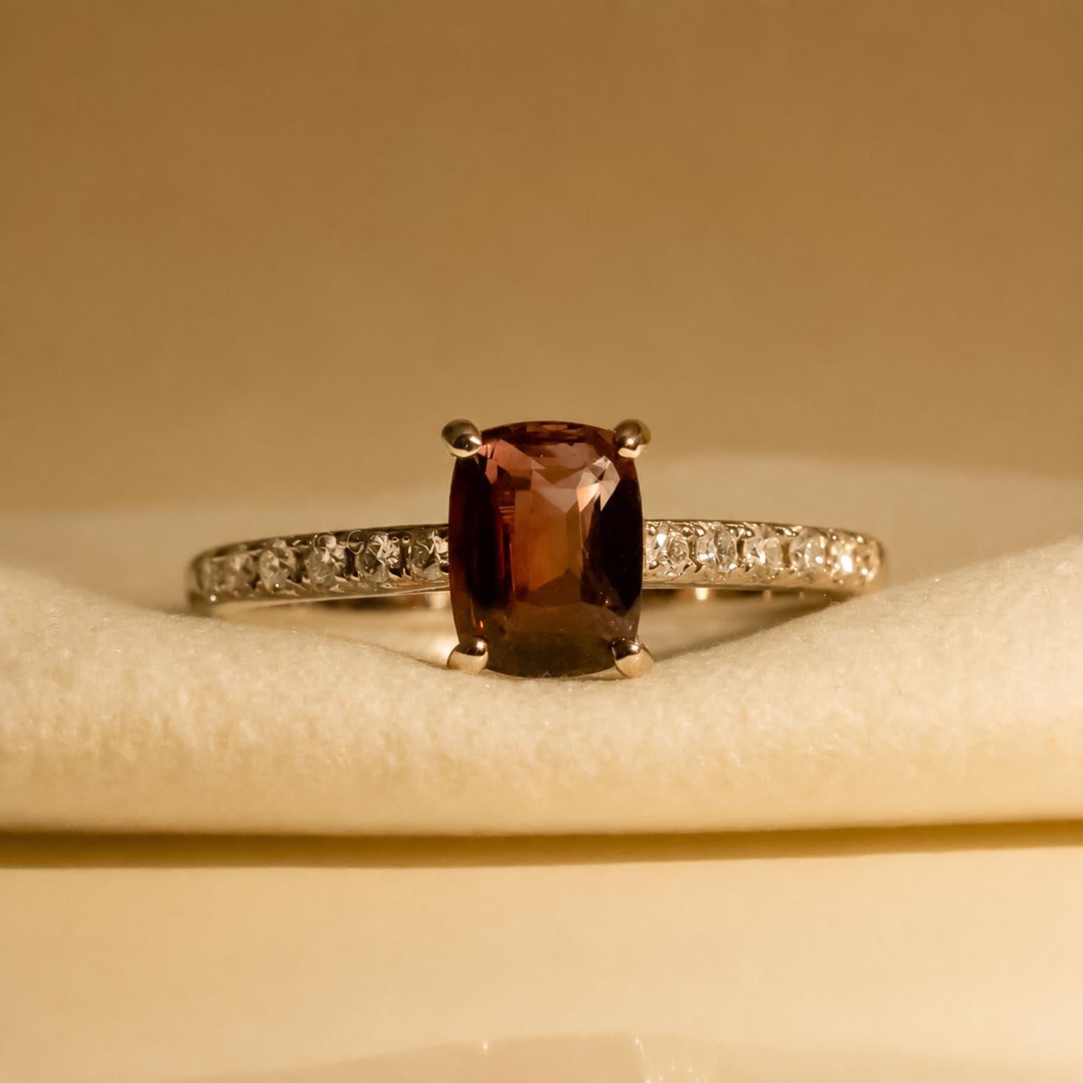A white gold ring featuring a cushion-cut alexandrite center stone and a pavé diamond band, displayed on a soft cream-colored velvet surface and shown under incandescent light 