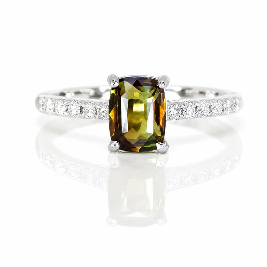 An 18K white gold diamond ring featuring a cushion-cut olive green alexandrite in a four-prong setting, with a pavé band of small diamonds, shown on a reflective white surface.