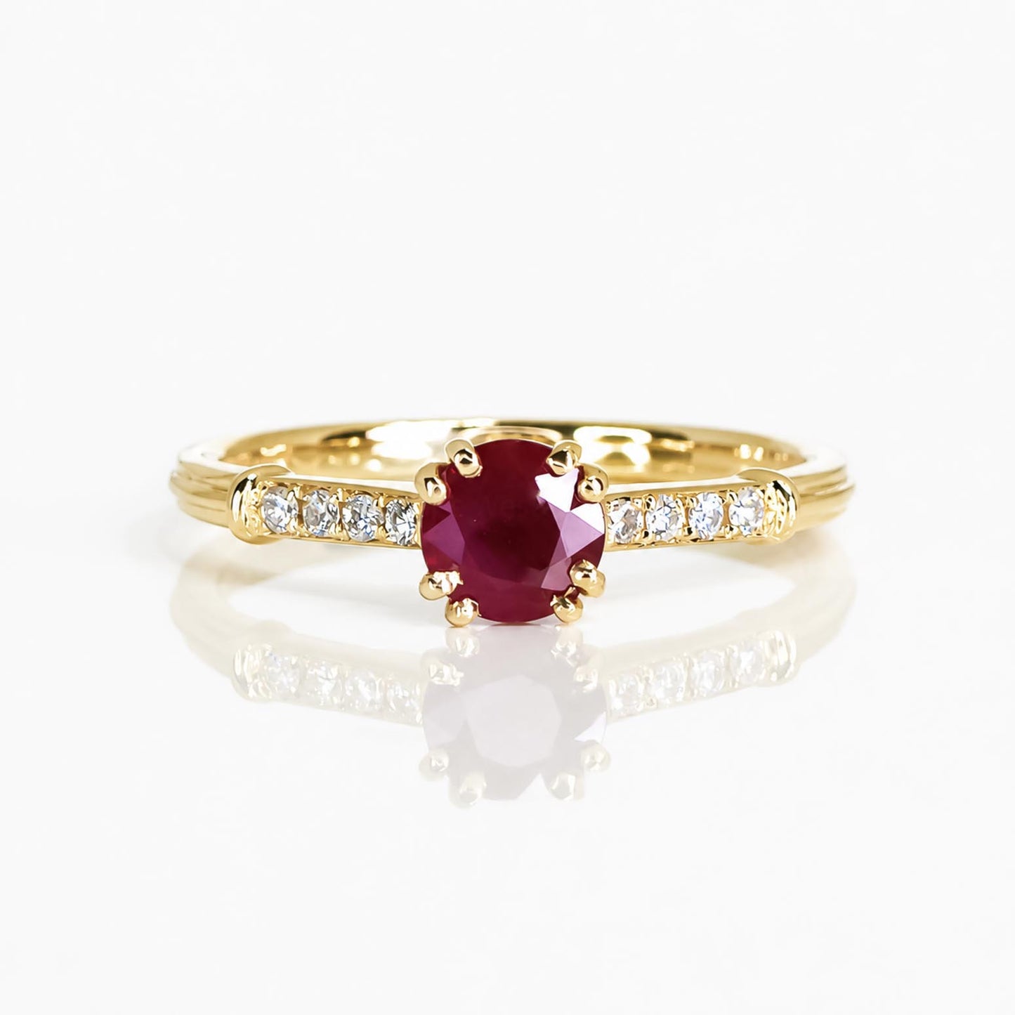 A yellow gold ring featuring a round-cut deep red ruby center stone, accented by small round-cut clear diamonds along the shoulders, set against a clean white background with a soft reflection.