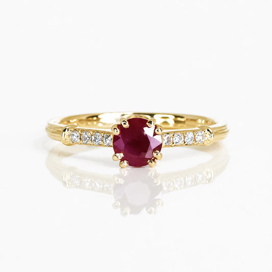 A yellow gold ring featuring a round-cut deep red ruby center stone, accented by small round-cut clear diamonds along the shoulders, set against a clean white background with a soft reflection.