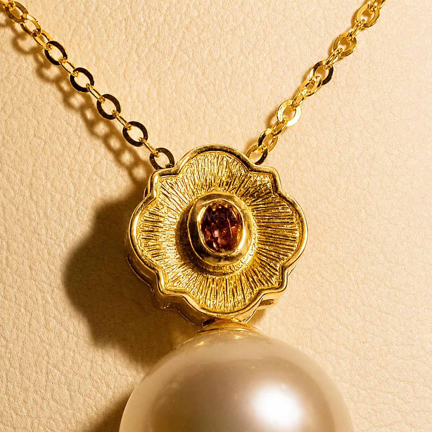 A gold pendant necklace featuring a textured flower-shaped setting with a bezel set oval alexandrite and a large, lustrous white pearl hanging below shown under incandescent light on a neutral background 