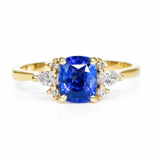 A blue sapphire ring with accent side diamonds in round and pear brilliant cut set in 18K yellow gold on a polished band photographed in white background