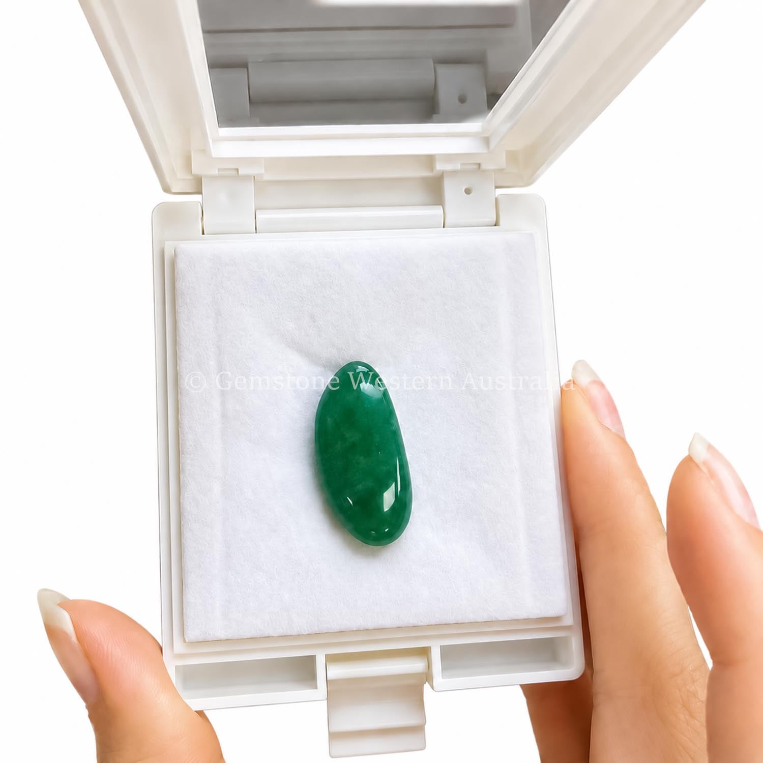 A polished Colombian emerald oval-shaped cabochon displayed in an open white presentation case with a white insert, held by two hands with manicured nails against a white background.