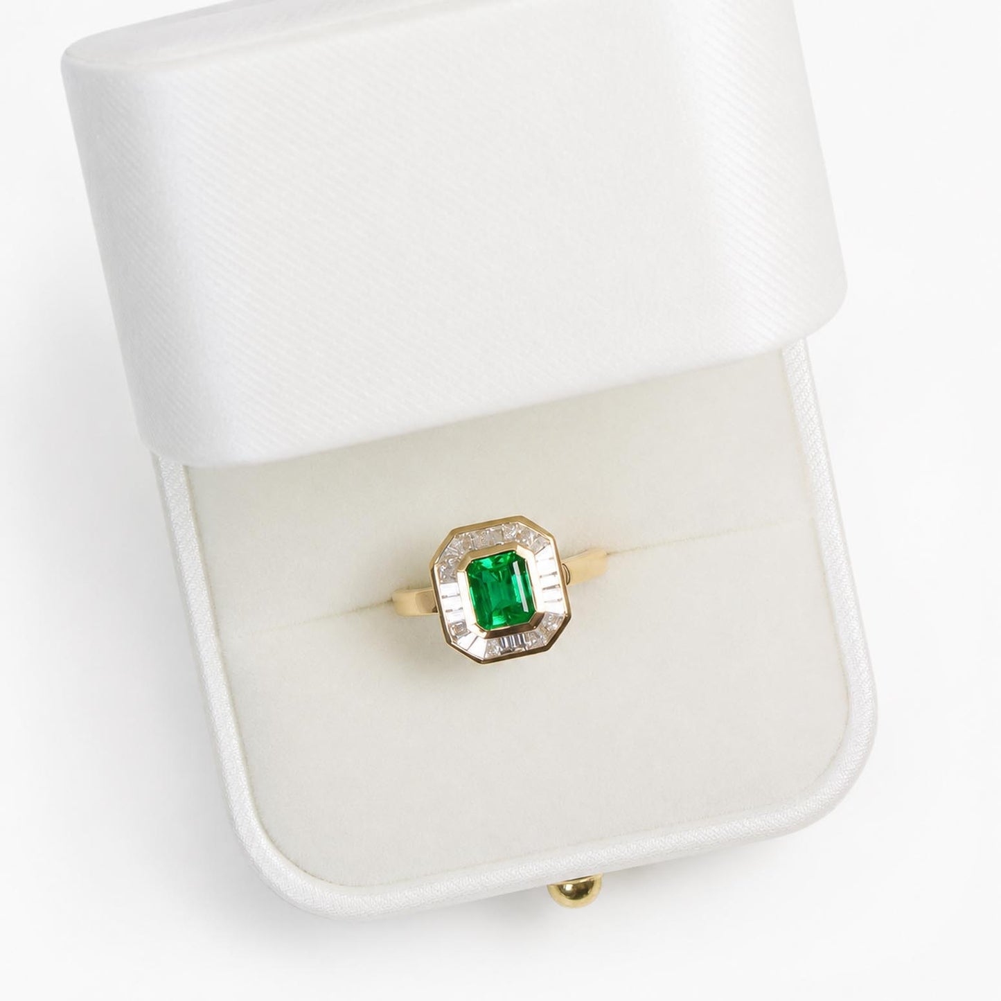 A yellow gold ring with a central emerald-cut Colombian emerald and a halo of baguette diamonds, displayed inside an open white satin ring box on a white background.