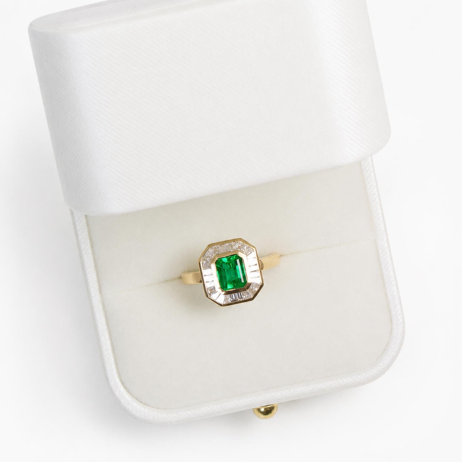 A yellow gold ring with a central emerald-cut Colombian emerald and a halo of baguette diamonds, displayed inside an open white satin ring box on a white background.