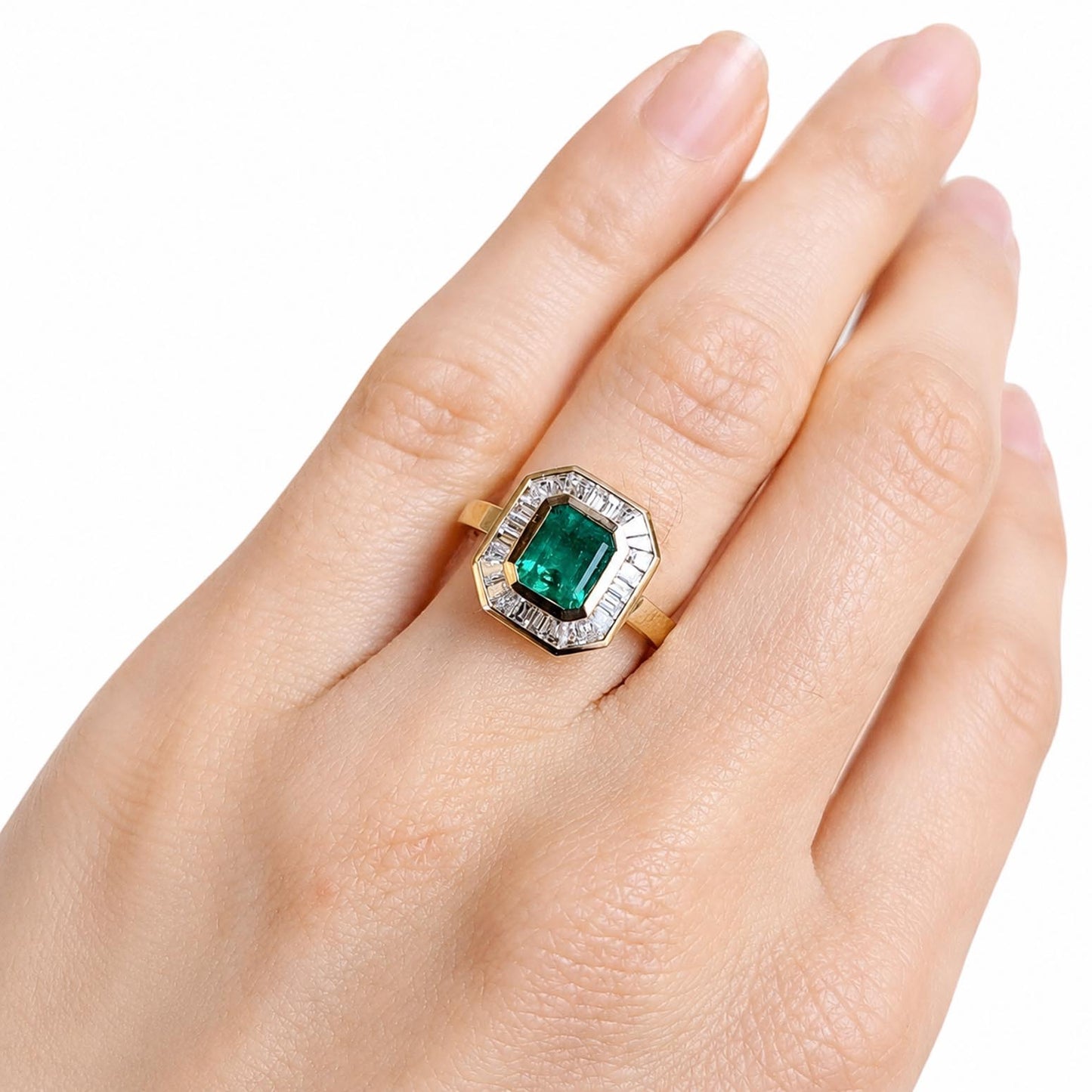 A yellow gold ring featuring an emerald-cut natural Colombian emerald surrounded by a halo of baguette diamonds, shown worn on a hand against a white background.