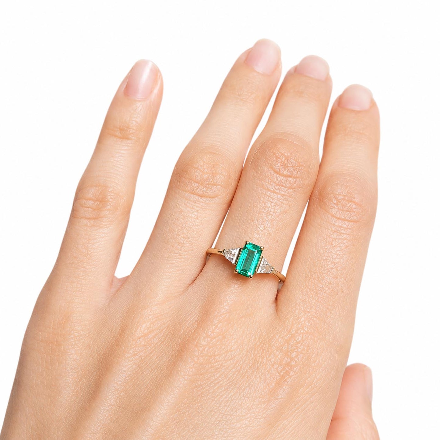 A close-up of a hand wearing a gold ring with a rectangular Colombian emerald and side diamonds set against a plain white background.