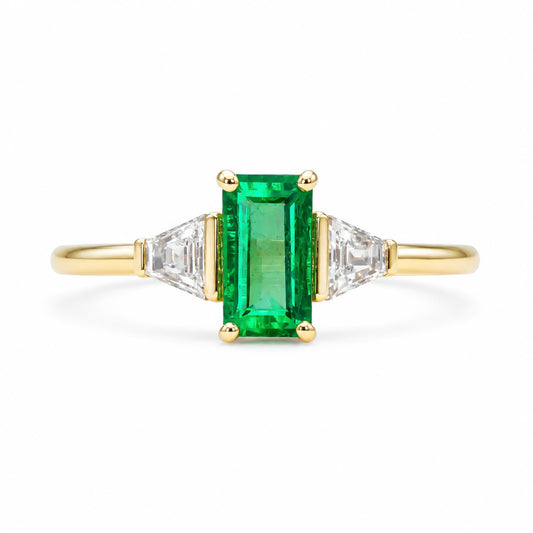A yellow gold ring featuring a rectangular emerald-cut Colombian emerald in the center, flanked by two tappered baguette-cut diamonds on a clean white background.