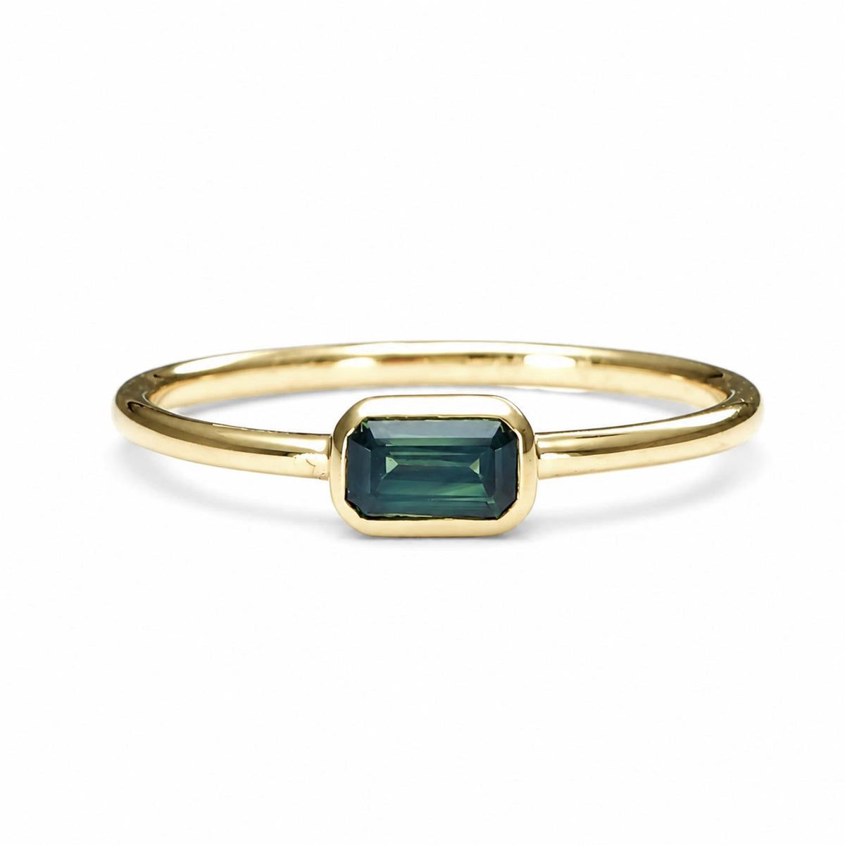 Yellow gold ring holding an Australian teal sapphire in emerald cut shape that is bezel set, shown on a white background
