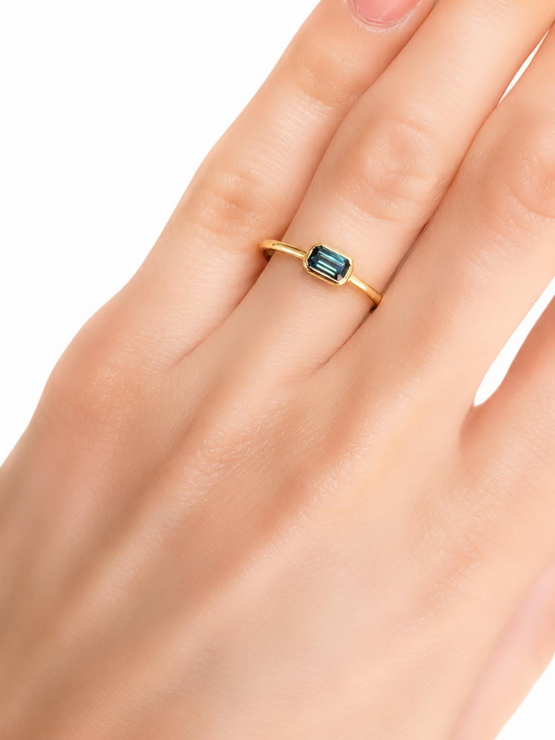 a hand wearing a Yellow gold ring holding an Australian teal sapphire in emerald cut shape that is bezel set, shown on a white background