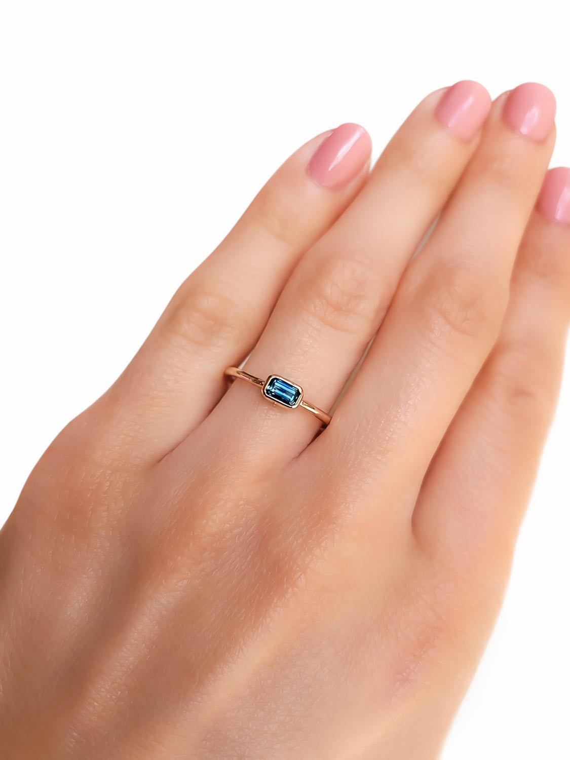 close up of a hand wearing a rose gold ring holding an Australian teal sapphire in emerald cut shape that is bezel set, shown on a white background