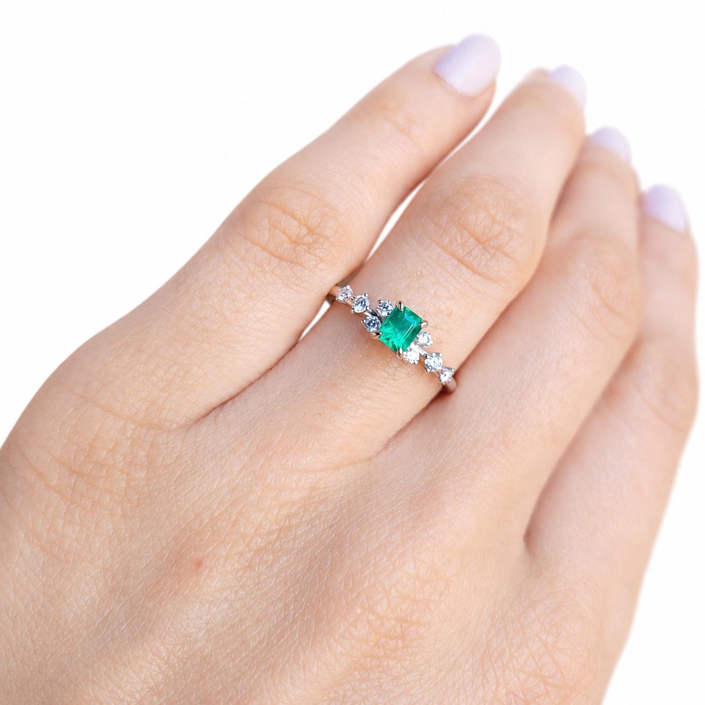 A close-up of a hand wearing a platinum ring with a central Colombian emerald and small diamond accents on either side, set against a plain white background.
