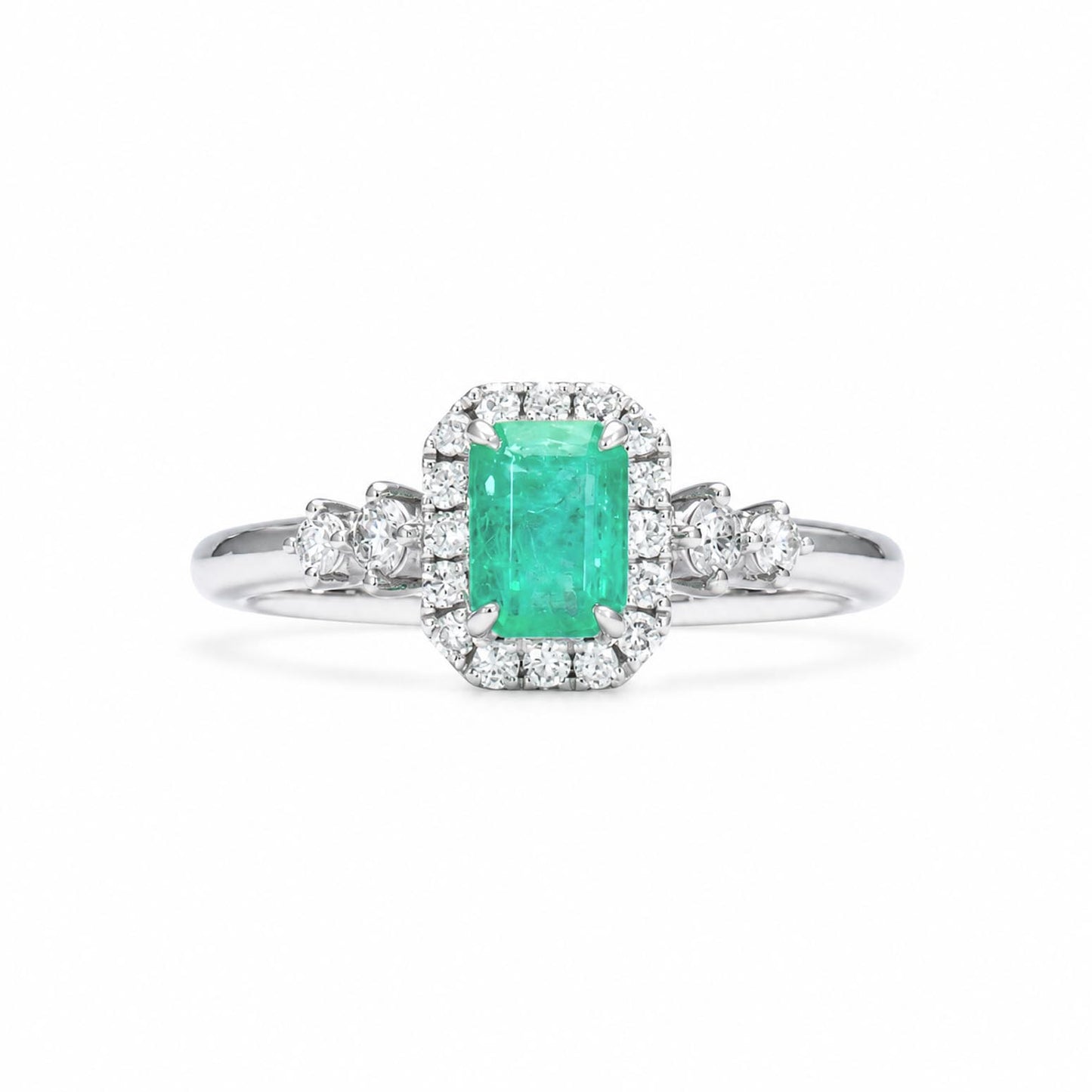 A Colombian emerald ring with an emerald-cut emerald surrounded by a diamond halo, diamond accents on the shank, shown on a clean white background.