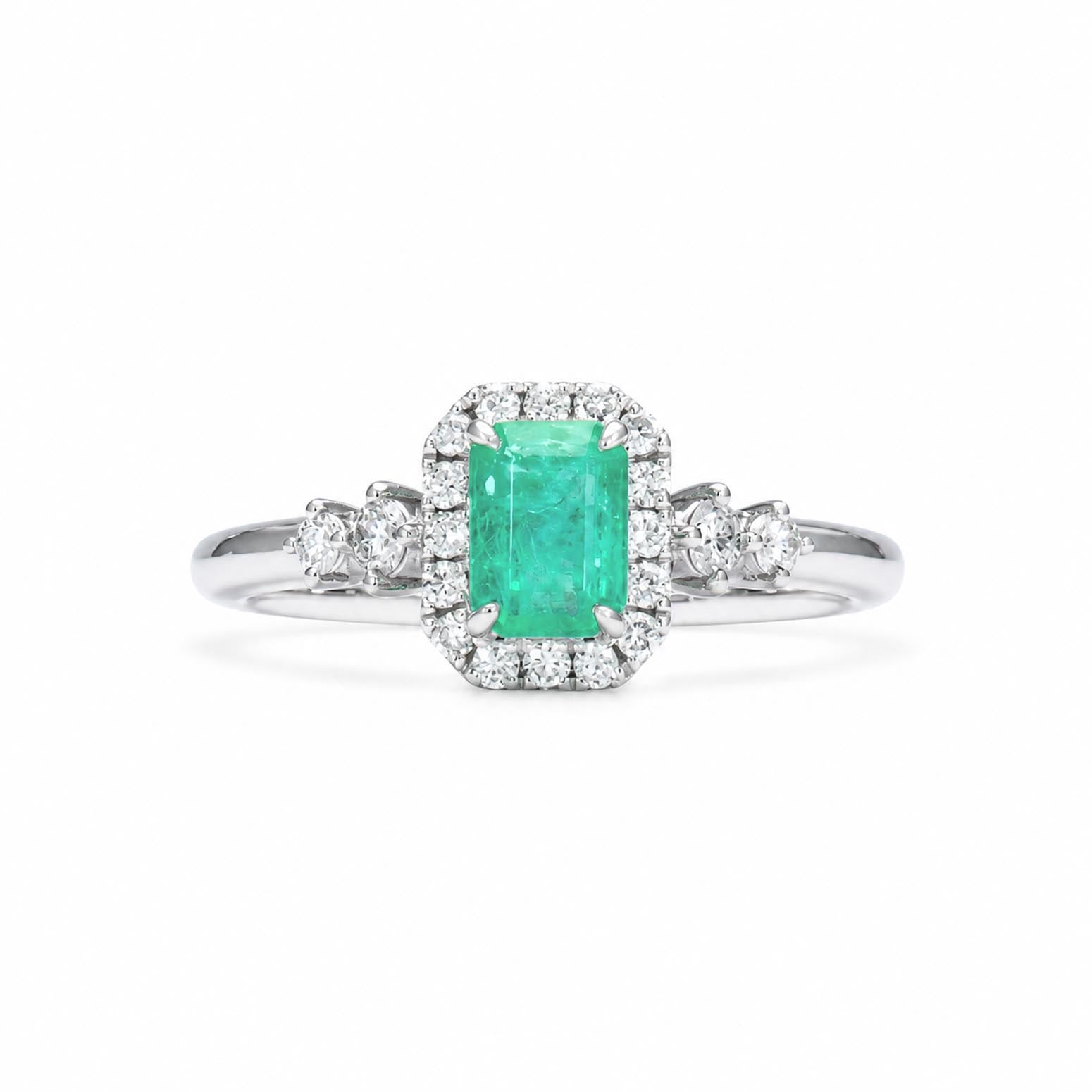 A Colombian emerald ring with an emerald-cut emerald surrounded by a diamond halo, diamond accents on the shank, shown on a clean white background.