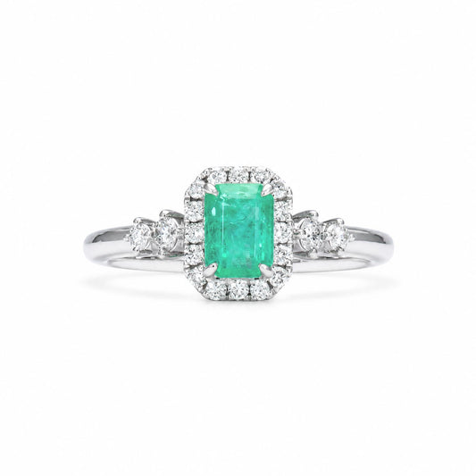 A Colombian emerald ring with an emerald-cut emerald surrounded by a diamond halo, diamond accents on the shank, shown on a clean white background.