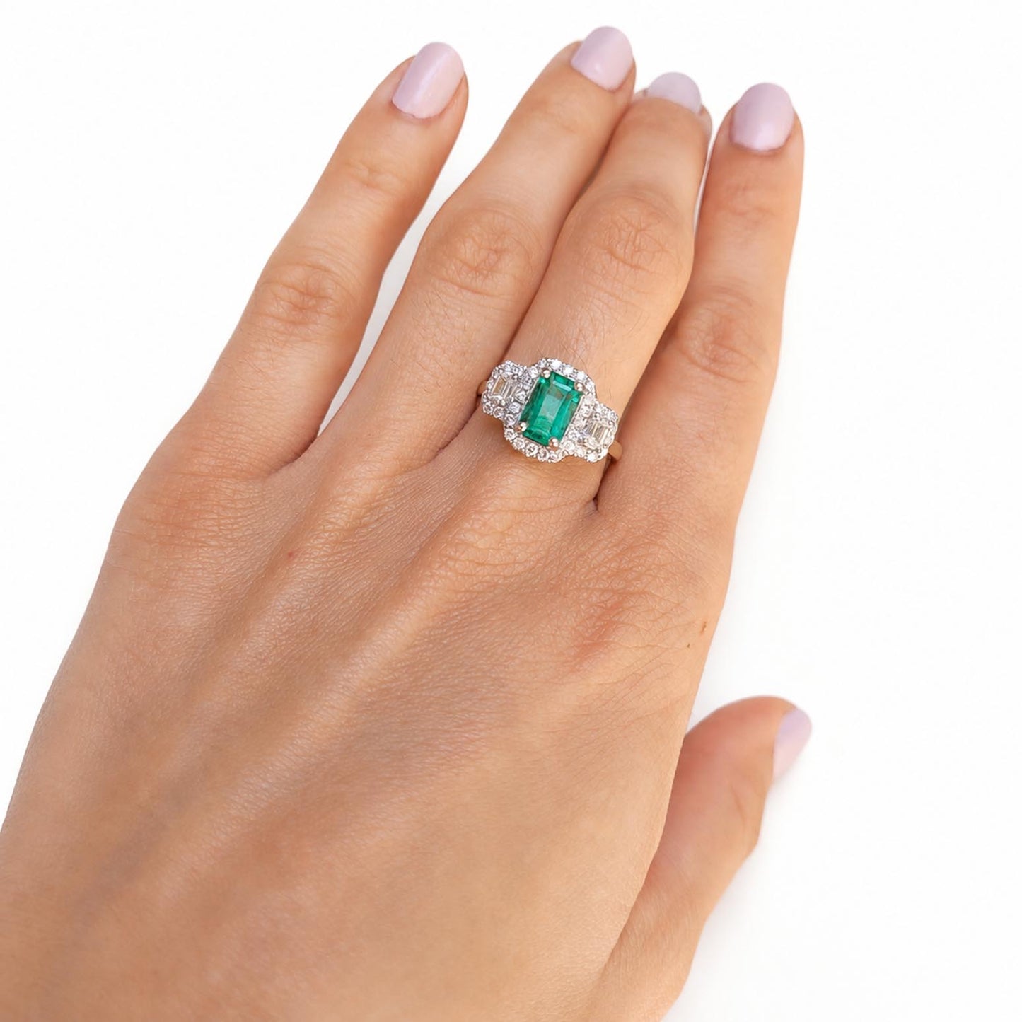 A close-up of a hand wearing a mixed metals platinum and yellow gold ring with an elongated Colombian emerald center stone and diamond accent stones, shown against a plain white background.